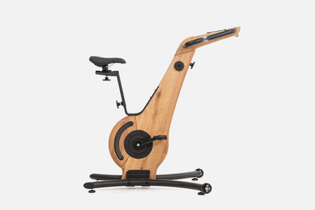 Side view of a pale wood indoor cycle with a black saddle, mesh flywheel cover, and a handlebar touchscreen