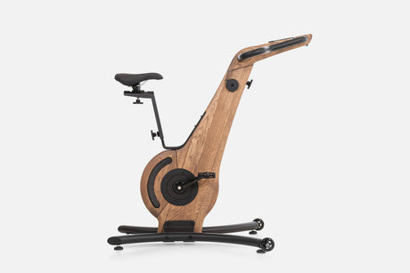 Side view of a medium-brown wooden indoor cycle with a black saddle, mesh flywheel cover, and a handlebar touchscreen