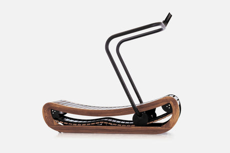 Side view of a curved manual treadmill with a medium-brown wood frame, black slats, and a raised black handlebar